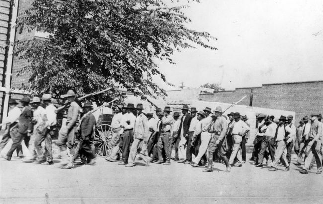 Grupė Nacionalinės gvardijos karių, nešiodama šautuvus su pritvirtintais durtuvais, palydi neginkluotus juodaodžius į sulaikymo centrą po Talsos lenktynių žudynių Talsoje, Oklahomoje, 1921 m. birželį.