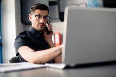 Vyras su akiniais telefone, naudodamas nešiojamąjį kompiuterį