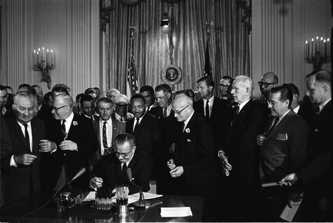Prezidentas Lyndonas B. Johnsonas pasirašo 1964 m. Civilinių teisių aktą, kai žiūri Martinas Lutheris Kingas jaunesnysis ir kiti.