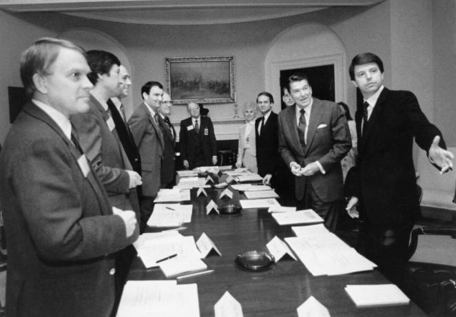 Nespalvotas Ronaldo Reagano ir kelių kitų kostiumuotų vyrų atvaizdas aplink ilgą konferencijų stalą
